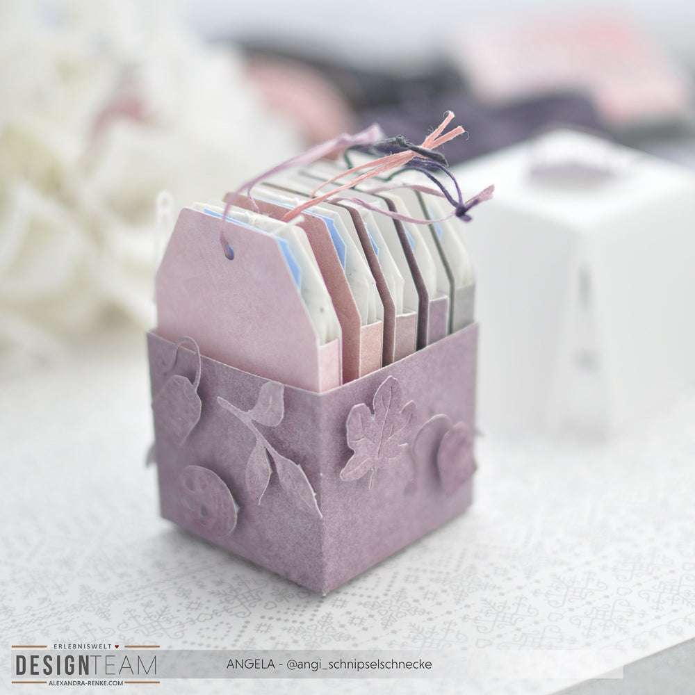Eine lilafarbene Papierschachtel von Alexandra Renke enthält pastellfarbene Geschenkanhänger mit Schnur und Blattverzierungen - perfekt zum Basteln mit Stanzen für Papier oder Ihrem DieCut System, um jeden Anhänger zu personalisieren.