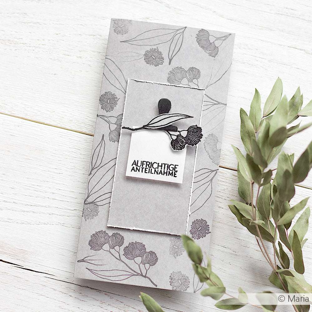 Graue Beileidskarte mit floralen Motiven von Alexandra Renke auf hochwertigem Designpapier, mit deutschem Text, neben grünen Blätterzweigen auf weißem Untergrund - ideal für besondere Papierprojekte.