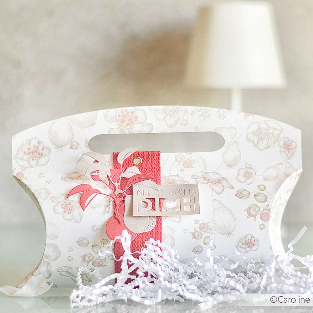 Dekorative, geblümte Geschenktüte von Alexandra Renke aus hochwertigem Designpapier mit rosa Akzenten und deutschem Etikett, abgebildet neben einer weißen Lampe und geschreddertem Papier - ideal für Fans von Papierprojekten oder Buchbinderei.