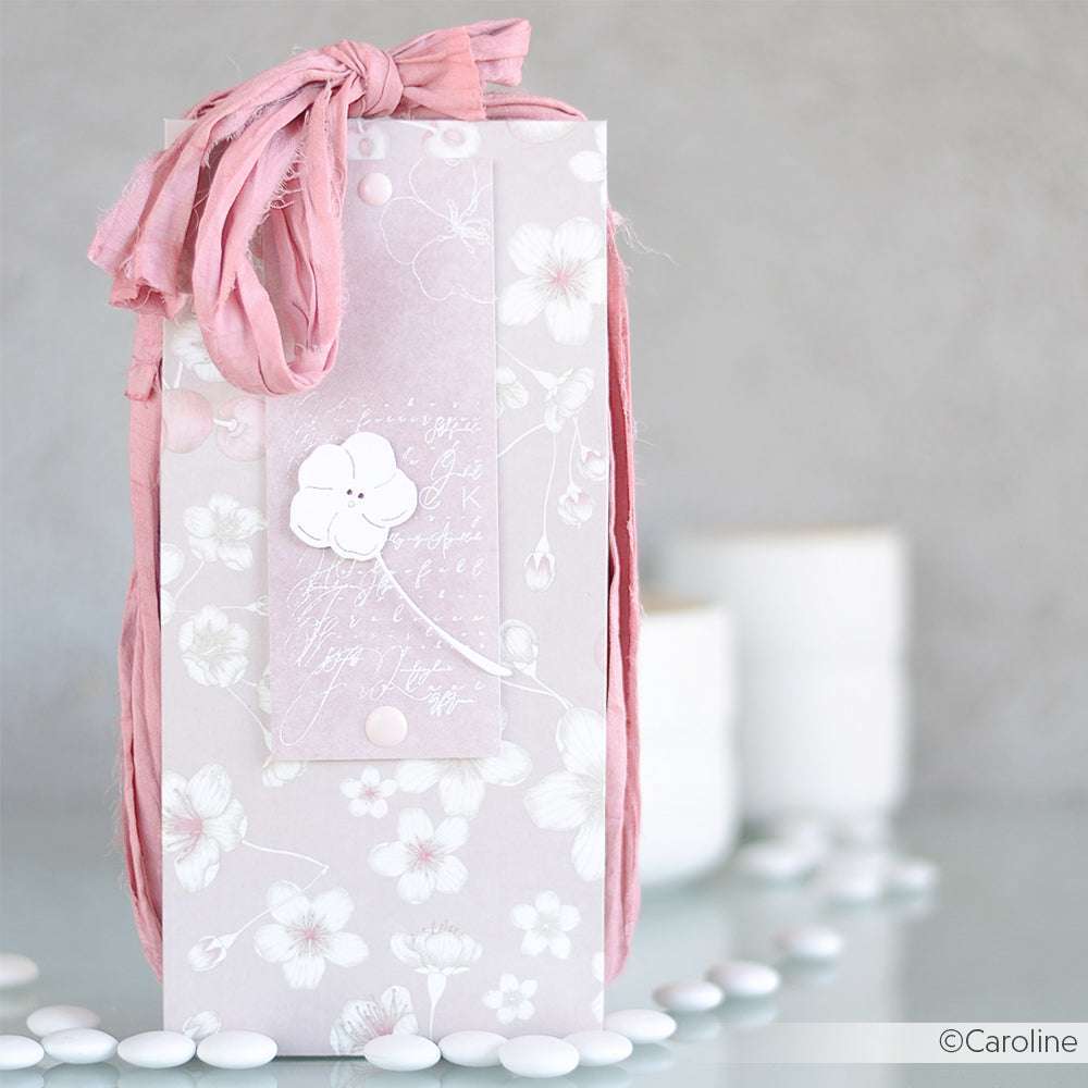 Blassrosa geblümte Geschenktüte aus Designpapier von Alexandra Renke, gebunden mit einem Band und umgeben von weißen Bonbons, mit sanft verschwommenen weißen Kerzen im Hintergrund.