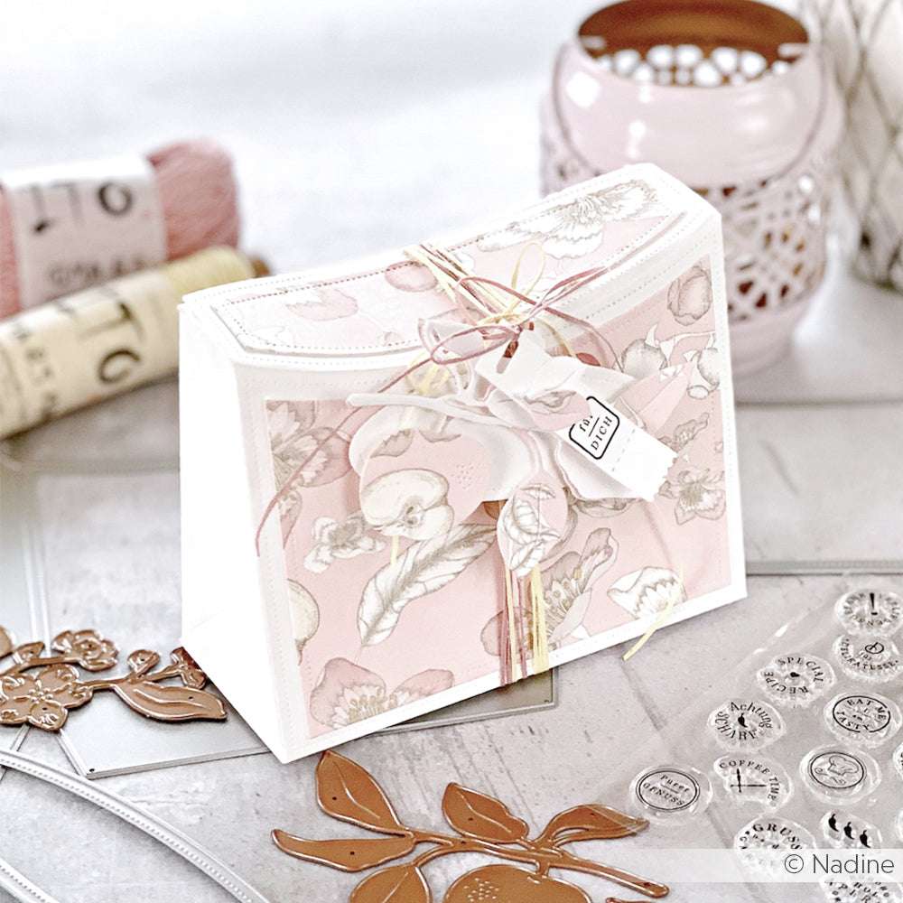 Eine florale Geschenkbox von Alexandra Renke mit einem Anhänger, umgeben von Bastelbedarf wie Stempeln, Stanzsystemen, Stanzformen, Bindfaden und rosa Garn.