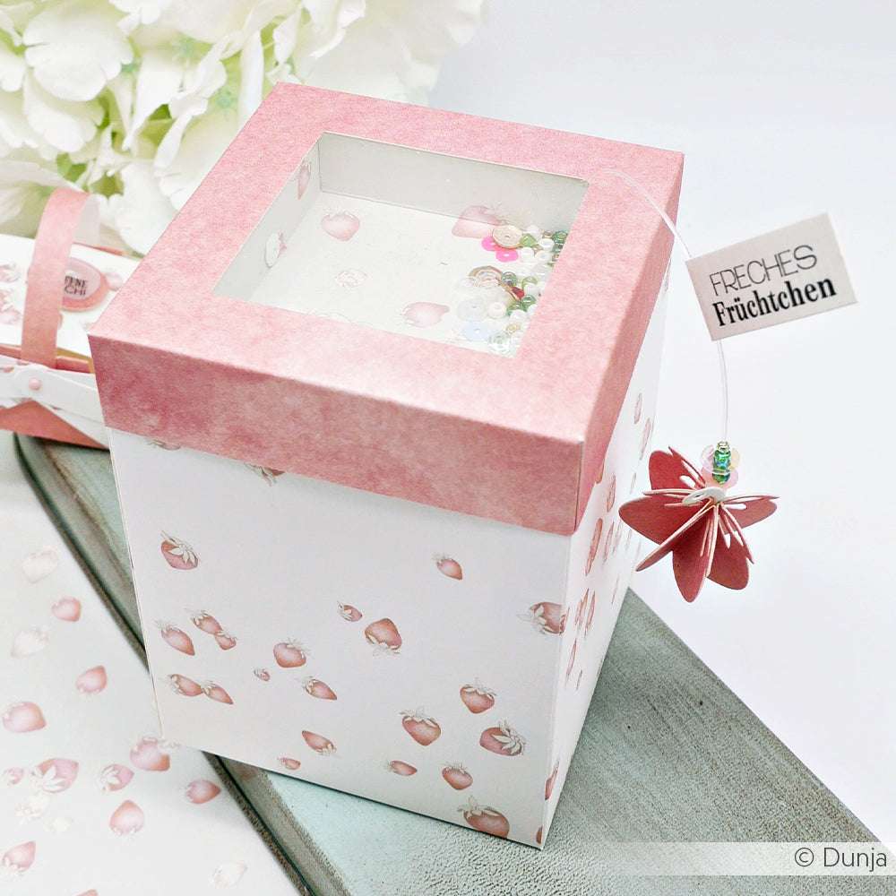 Eine rosa-weiße Alexandra Renke-Schachtel mit Erdbeeren, durchsichtigem Deckel und einem Blumenanhänger mit der Aufschrift "Freches Früchtchen" - ideal für Papierprojekte oder als Inspiration für Grußkarten mit dem Designpapier Mimi Dahlienrosa.