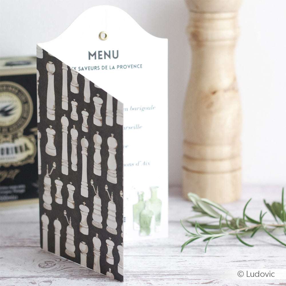 Eine elegante französische Menükarte von Alexandra Renke aus Designpapier steht auf einem Tisch, begleitet von einer Pfeffermühle, einer Schachtel und einem Rosmarinzweig.