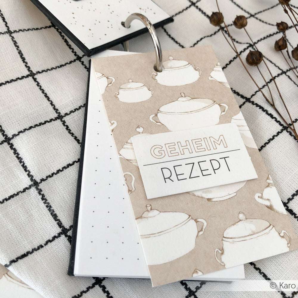 Die Rezeptkarten am Ring von Alexandra Renke, mit elegantem Designpapier und "Geheim Rezept" auf dem Umschlag, liegen auf einem karierten Tuch - ideal für Ihre kreativen Papierprojekte.