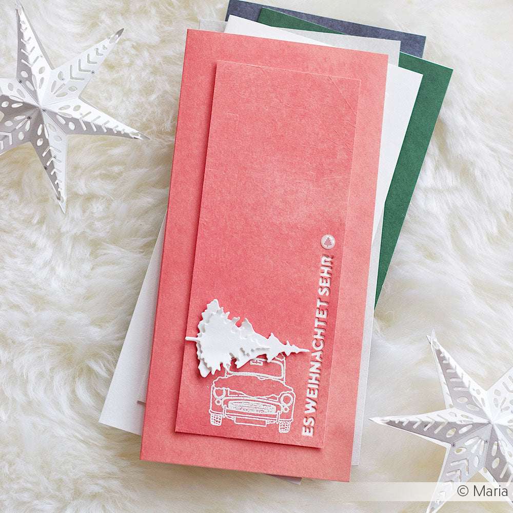 Rosa Weihnachtskarte mit einem Auto und einem Baum, entworfen von Mimi Fruchtrot für Alexandra Renke, umgeben von Papiersternen aus Designpapier auf einer weichen weißen Oberfläche.