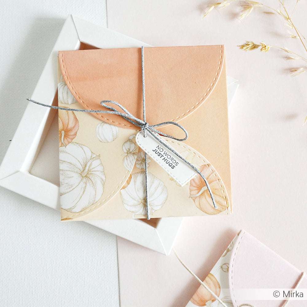 Quadratischer Umschlag mit floralem Muster aus Alexandra Renke Designpapier, verschnürt mit einer Schnur und einem Anhänger mit der Aufschrift "No words, just hugs" auf pastellfarbenem Hintergrund - ideal für einzigartige Papierprojekte.