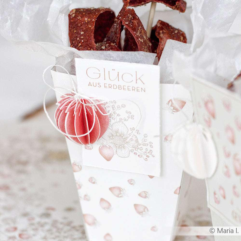 Weißer Geschenkkarton mit Erdbeerdruck, gefertigt aus Alexandra Renke Designpapier Erdbeer-Holunder und beschriftet mit Glück aus Erdbeeren. Verziert mit einem roten Papierornament - perfekt für Papierprojekte und zum Befüllen mit Leckereien.