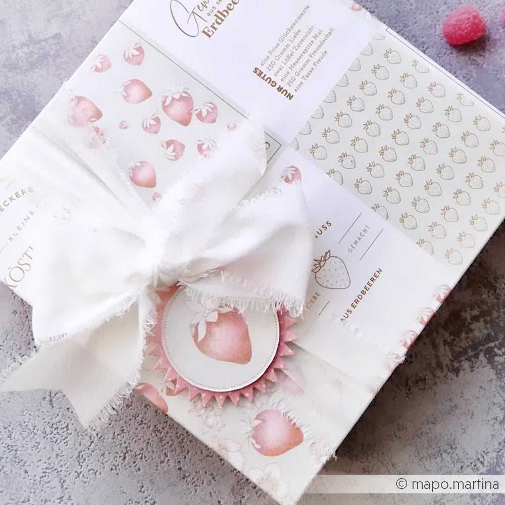 Geschenkkarton verpackt in Alexandra Renkes erdbeergemustertem Designpapier mit weißem Band und Erdbeeranhänger - perfekt für Papierprojekte oder zusammen mit dem Kärtchenbogen Erdbeer-Holunder auf einer hellen Fläche.