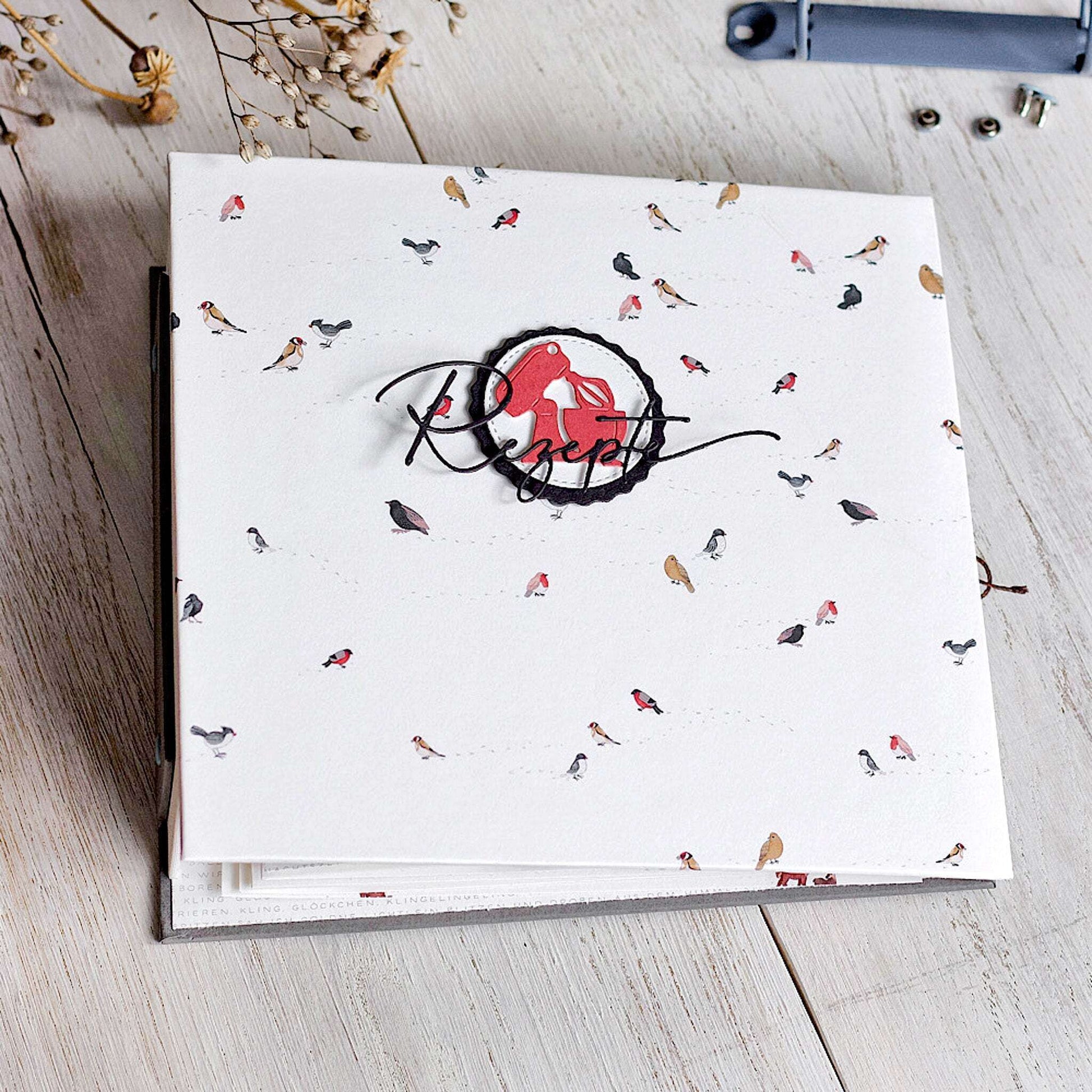 Ein Rezeptbuch von Alexandra Renke mit einem Einband aus Designpapier mit Vogelmotiven und "Rezepten" in Schreibschrift, ausgestellt auf einem Holztisch.