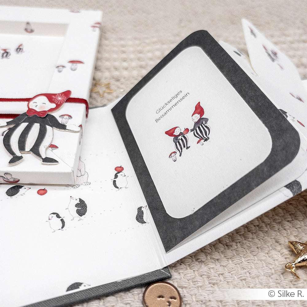 Eine offene handgefertigte Karte von Alexandra Renke mit skurrilen Clown-Illustrationen auf Designpapier ruht auf einer strukturierten beigen Oberfläche.