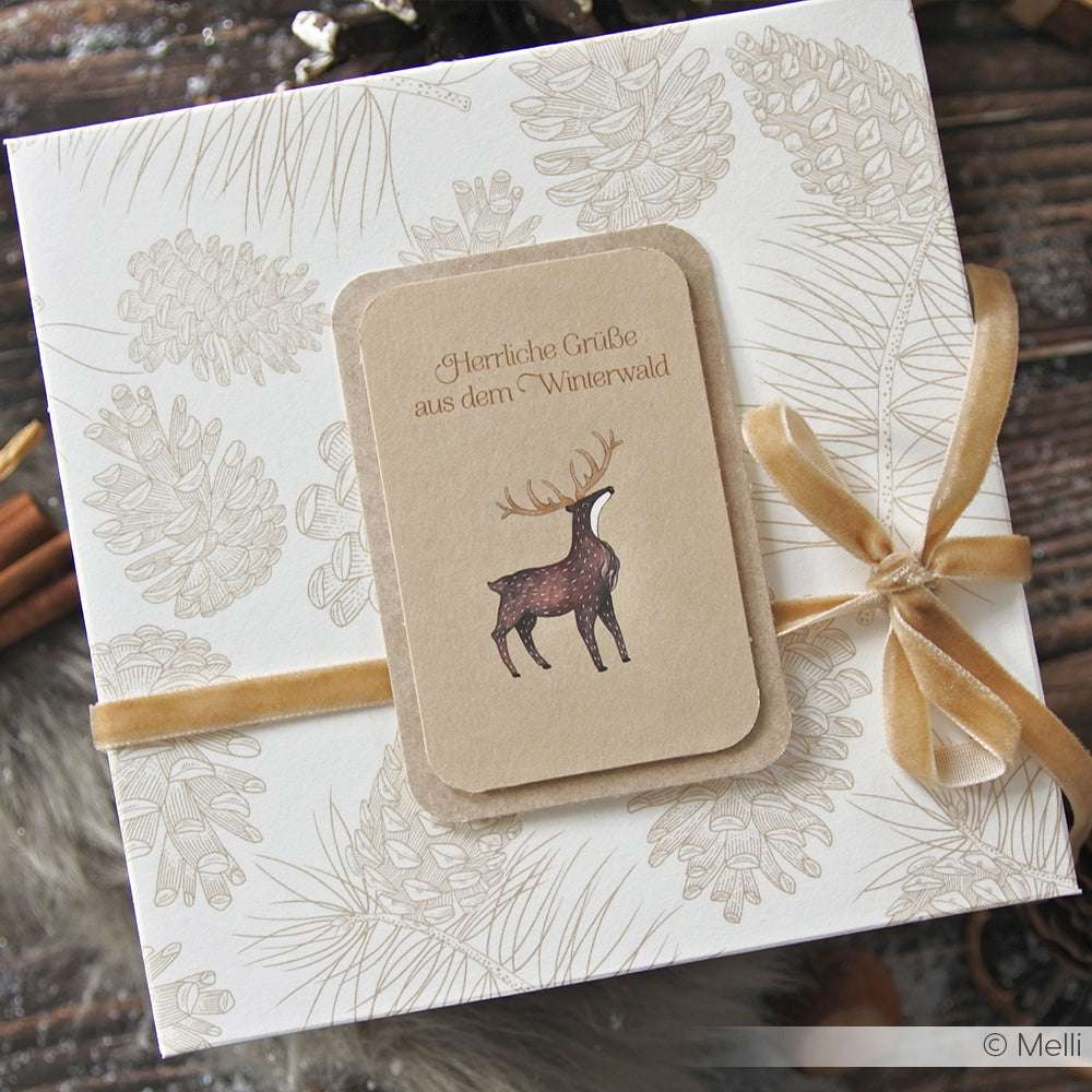 Geschenkkarton aus Alexandra Renke Tannenzapfen-Vielfalt Designpapier, veredelt mit einem hellbraunen Band und einer Hirschkarte mit einem deutschen Winterwaldgruß - ideal für festliche Papierprojekte.