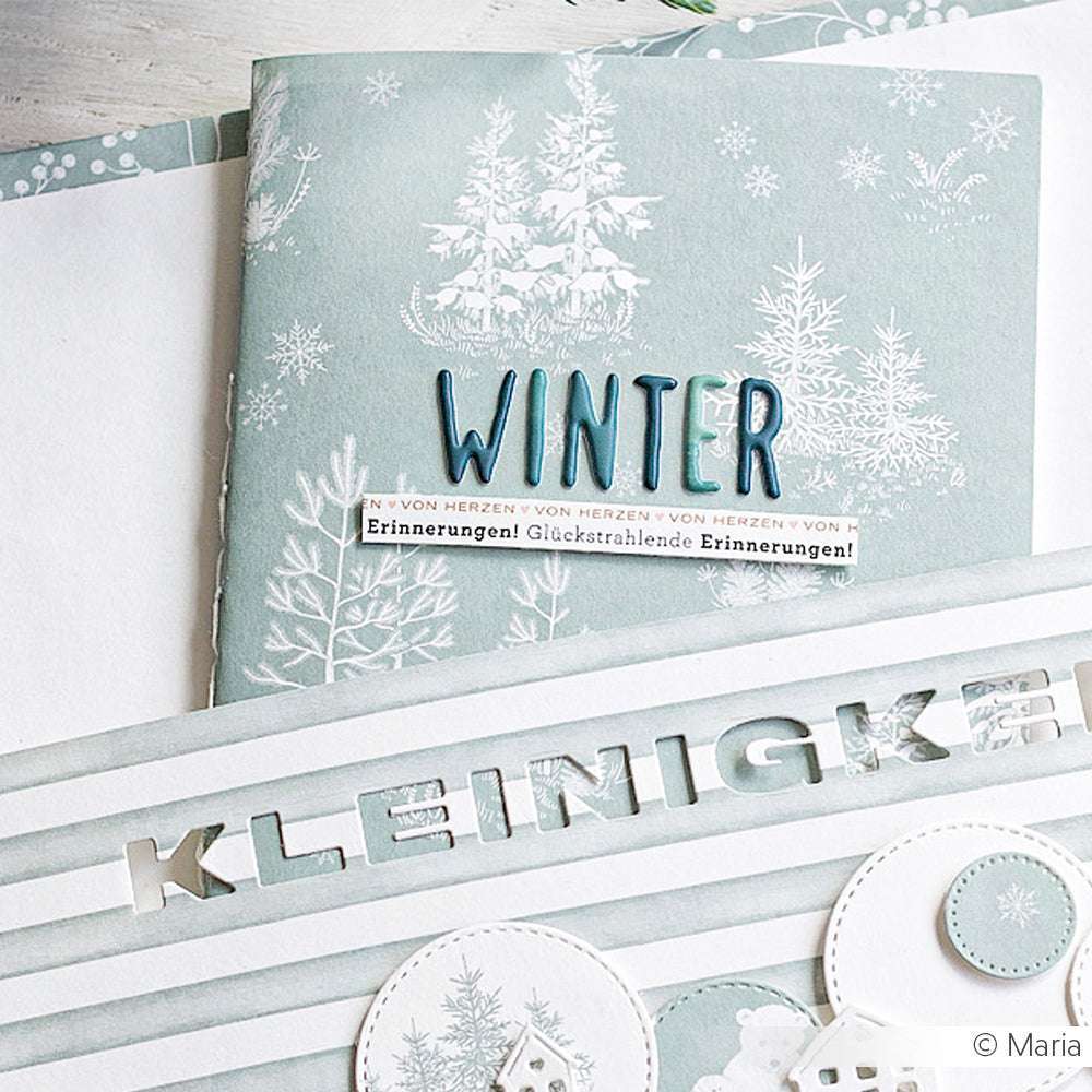 Das winterliche Scrapbook von Alexandra Renke zeigt "WINTER" und "KLEINIGKEITEN" auf blau-weißem Designpapier mit Baummotiven, die von einem verschneiten Wald inspiriert sind - ideal für Ihre Papierbastelprojekte.