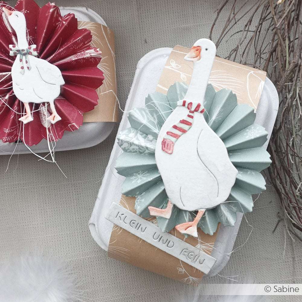 Geschenkschachteln von Alexandra Renke mit Akzenten von Jaspisgrün, Designpapier, Papierrosetten und ausgeschnittenen Gänsen in Schals vor neutralem Hintergrund.