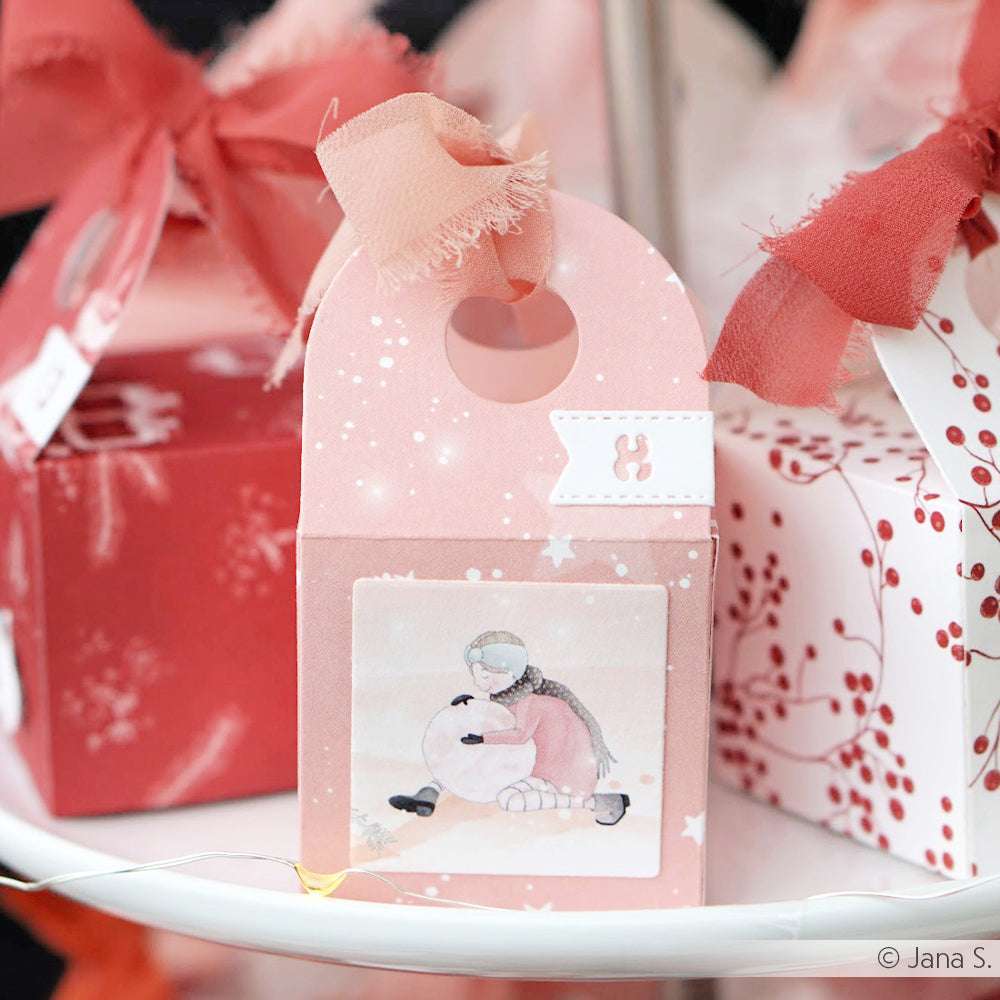 Rosafarbene Geschenkschachtel mit Schleife von Alexandra Renke mit einer Mädchen- und Schneemann-Illustration - ideal für Papierprojekte oder als Inspiration für liebevoll gestaltete Grußkarten auf Festtagsdisplays.