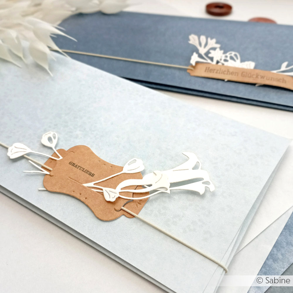 Die eleganten blauen Grußkarten von Alexandra Renke, die mit Designpapier erstellt wurden, enthalten weiße Blumenausschnitte und braune Etiketten mit deutschen Botschaften, die auf einer weißen Oberfläche angebracht sind.