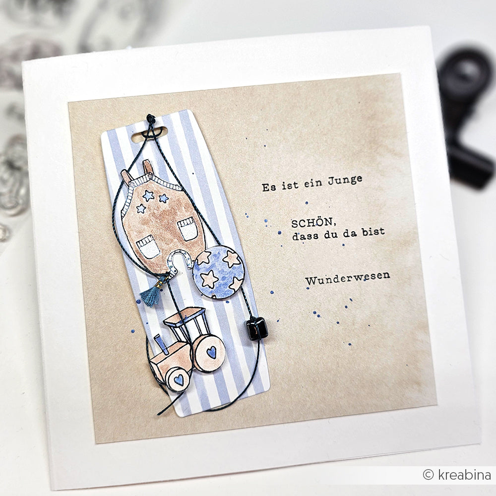 Handgemachte Karte von Alexandra Renke mit einem Haus-, Ballon- und Spielzeugdesign auf hochwertigem Designpapier mit deutschem Text zur Begrüßung eines kleinen Jungen.