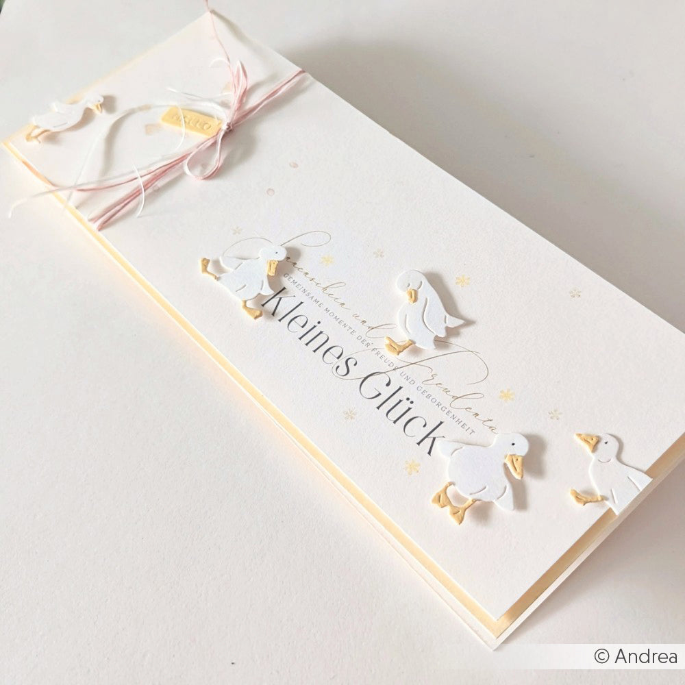 Eine weiße Karte von Alexandra Renke mit Designpapier, geprägten Enten, goldenen Akzenten und rosafarbener Kordel, mit "Meines Glück" auf der Vorderseite.