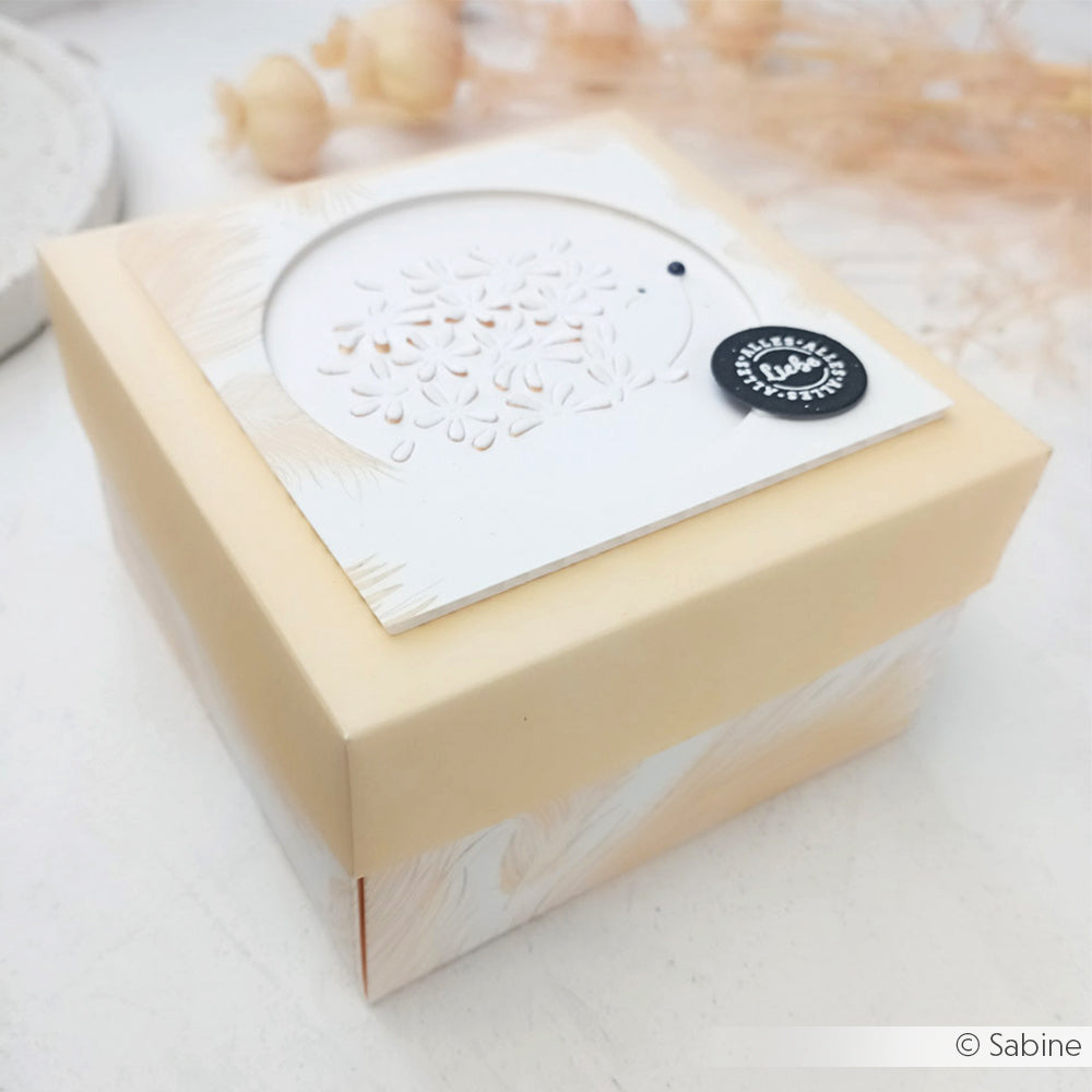 Eine cremefarbene Geschenkbox von Alexandra Renke mit floralem Deckelausschnitt, gefertigt aus hochwertigem Designpapier und mit einem kleinen runden schwarzen Anhänger versehen.