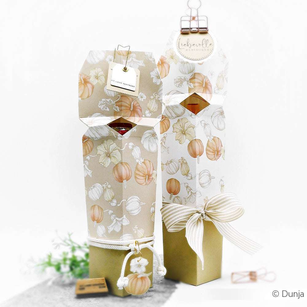 Zwei elegante Alexandra Renke Geschenkboxen mit herbstlichen Kürbismustern, hergestellt aus hochwertigem Designpapier und mit weiß-goldenen Bändern verschnürt, stehen auf weißem Hintergrund - perfekt für Papierprojekte oder Buchbinderei.