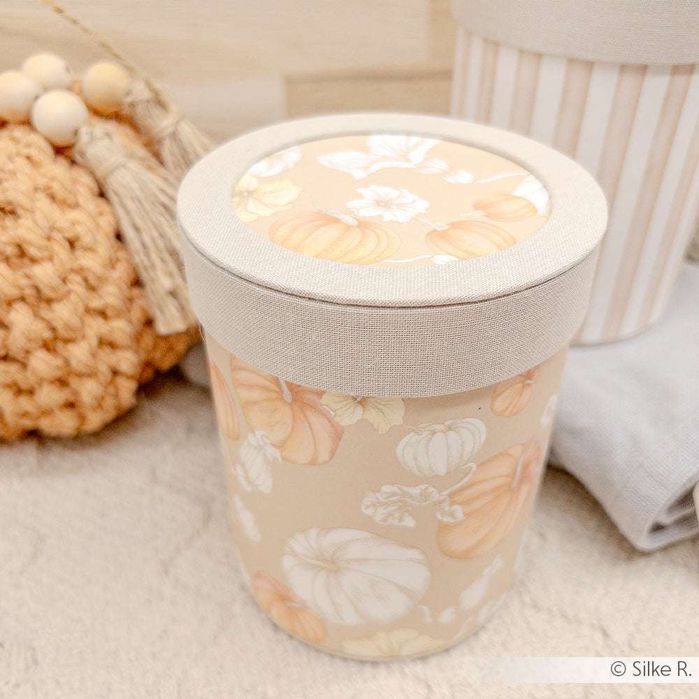 Ein beiger Behälter mit Kürbismuster von Alexandra Renke, ideal für Papierprojekte oder Buchbinderei, ruht auf einer hellen Stofffläche neben einem gehäkelten Gegenstand.