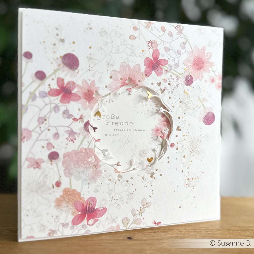 Quadratische Blumenkarte aus Alexandra Renke Designpapier mit rosafarbenen Blumen, einem runden Ausschnitt und deutschem Text auf einer Holzoberfläche. Ideal für Papierprojekte oder Buchbinderei.