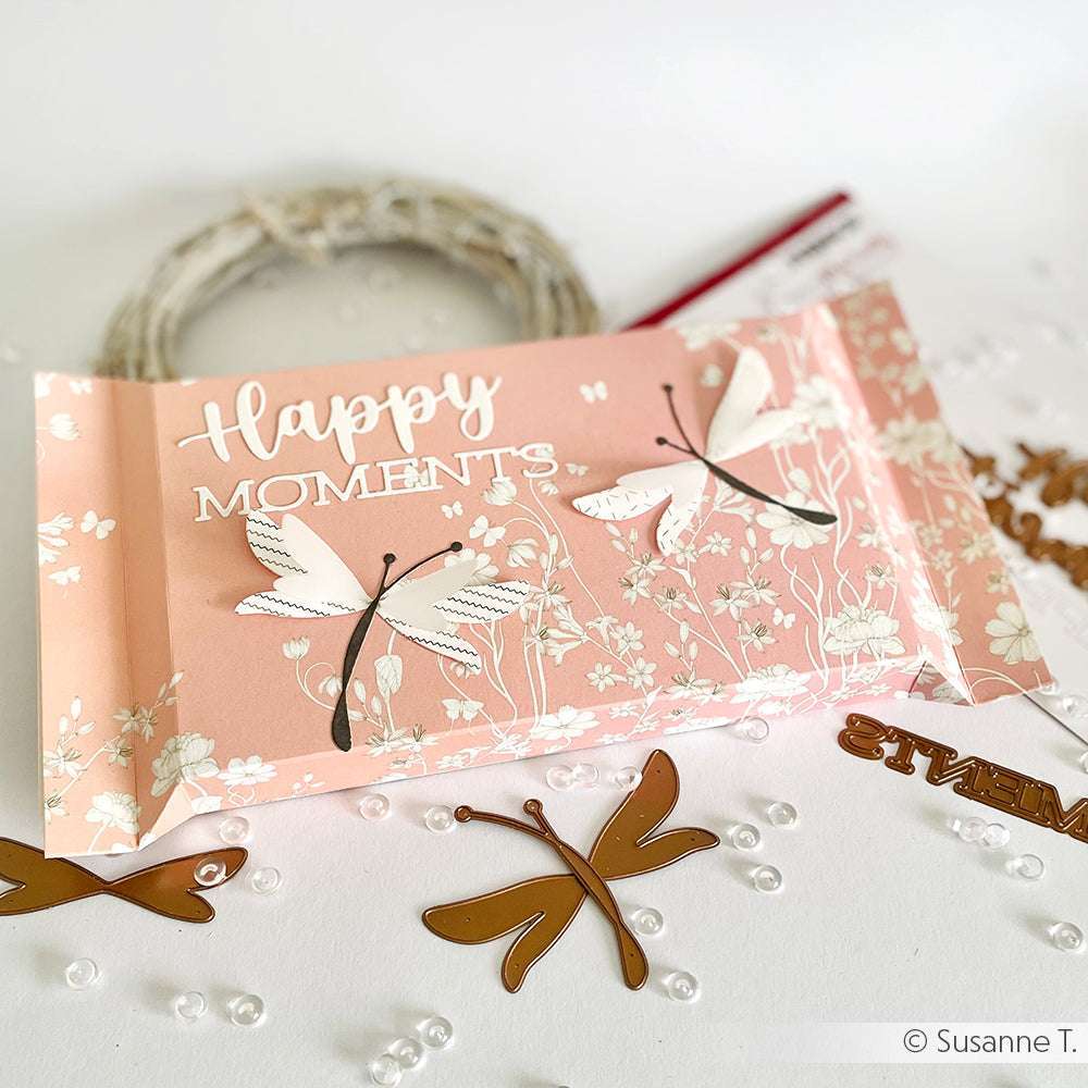 Eine rosafarbene Karte mit "Happy Moments" und Libellenmustern, hergestellt mit Alexandra Renke Designpapier, verziert mit Perlen und goldenen Libellenausschnitten - ideal für Papierbastelprojekte.