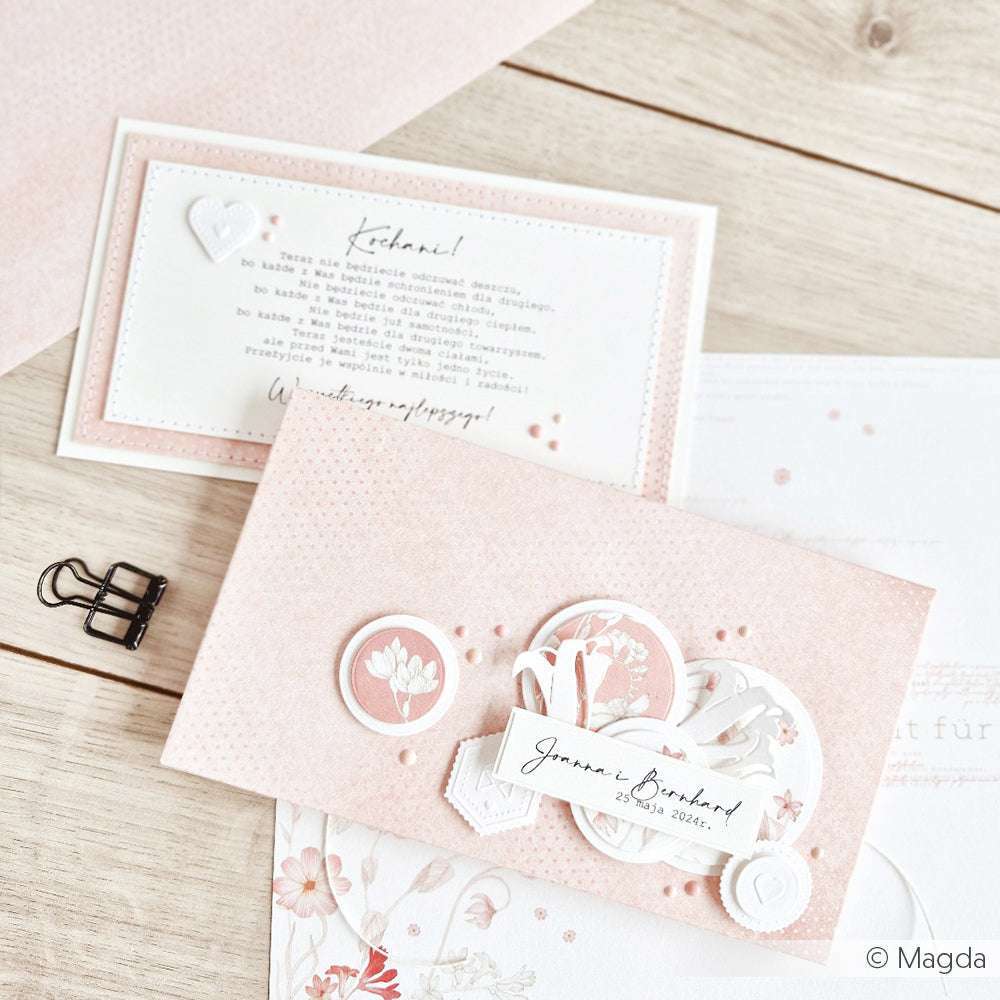 Eine rosa-weiß geblümte Hochzeitseinladung von Alexandra Renke mit dekorativen Designpapierkarten auf einer Holzoberfläche.