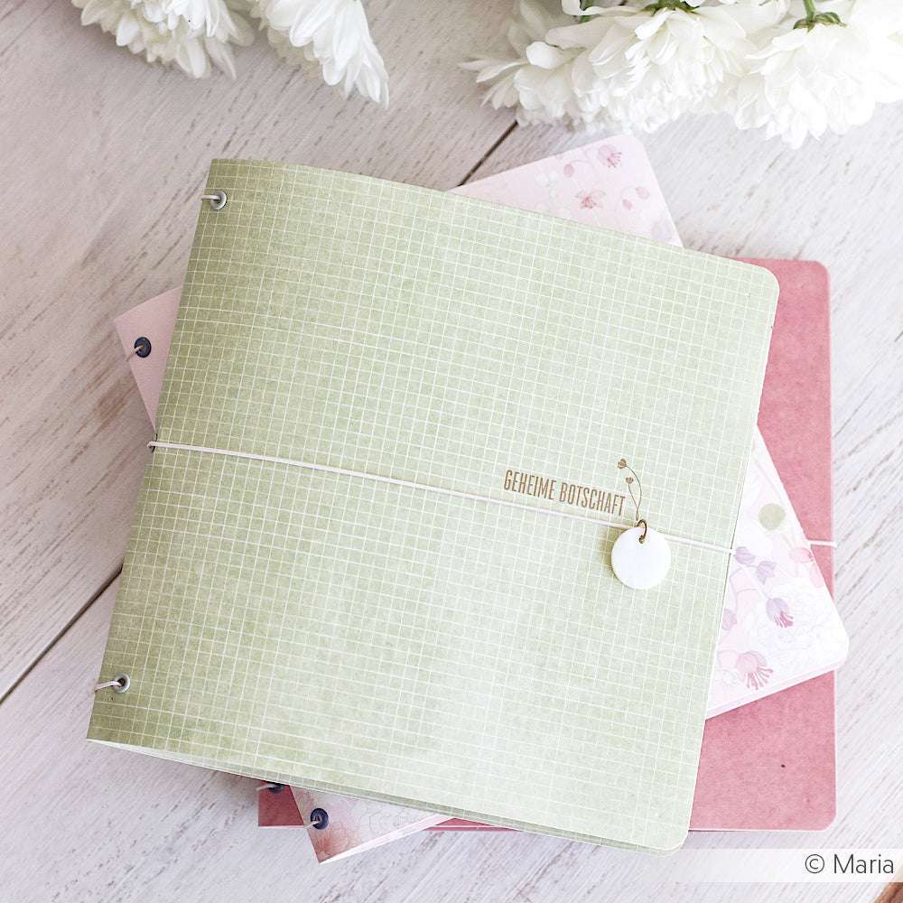 Ein grünes Notizbuch mit Gittermuster von Alexandra Renke liegt auf zwei pastellfarbenen Notizbüchern, daneben stehen weiße Blumen.