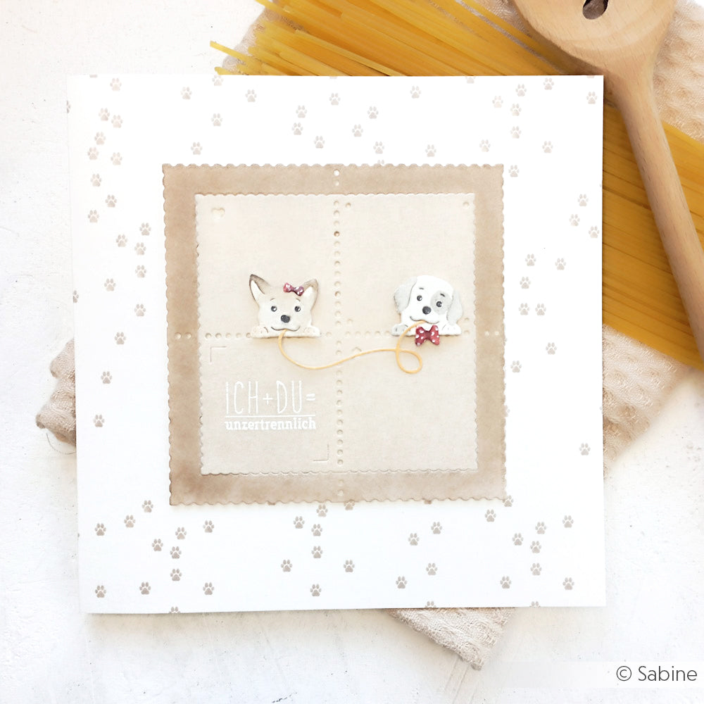 Die Grußkarte von Alexandra Renke auf Designpapier zeigt zwei Cartoon-Hunde, die mit einer Schnur verbunden sind, umgeben von Pfotenabdrücken und Kochutensilien - ideal für Papierprojekte oder als Inspiration für die Buchbinderei.