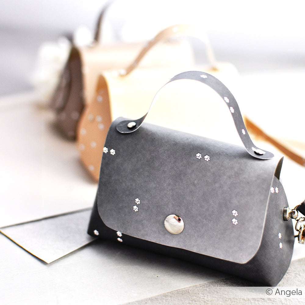 Eine kleine graue Handtasche von Alexandra Renke mit einem Henkel, einem silbernen Knopf und winzigen weißen Blumenmustern - ideal für Papierprojekte oder inspiriert von der Buchbinderei.