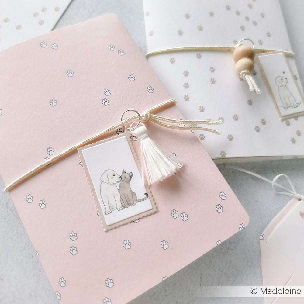 Zartrosa und weiße Notizbücher mit Pfotenabdrücken und Hund-Katzen-Anhängern, gebunden mit Schleife und Quaste - perfekt für Papierprojekte oder Buchbinderei von Alexandra Renke.