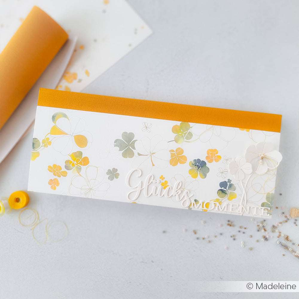 Eine florale Karte von Alexandra Renke mit orangefarbenem Rand, Kleeblättern und "Glücksmomente" auf einer hellen Oberfläche mit Designpapier - ideal für Papierprojekte oder Buchbinderei.