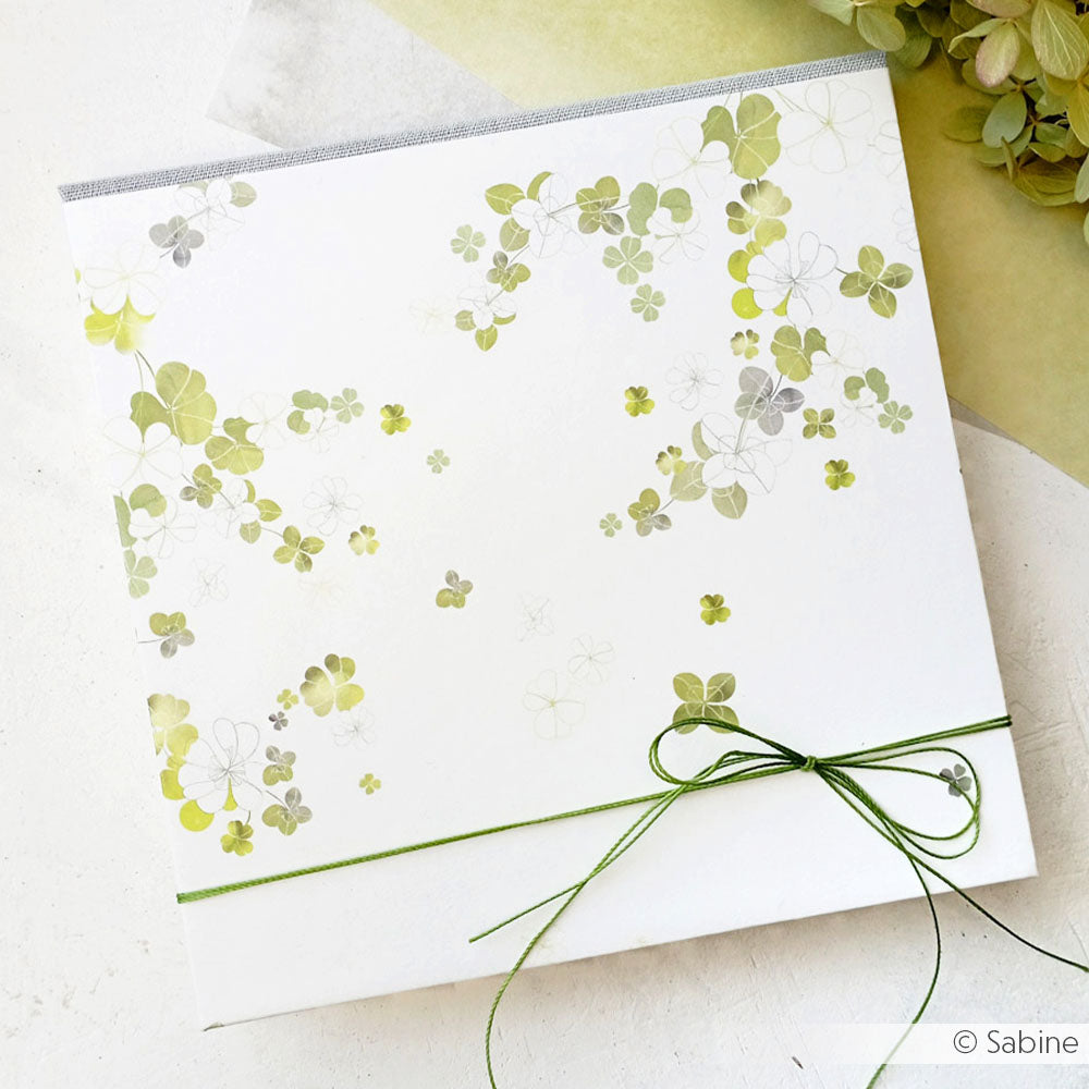 Quadratische Karte von Alexandra Renke mit zartem Grünem Glücksklee-Gestöber und einer grünen Kordelschleife in der rechten unteren Ecke - ideal für Designpapier Papierprojekte.