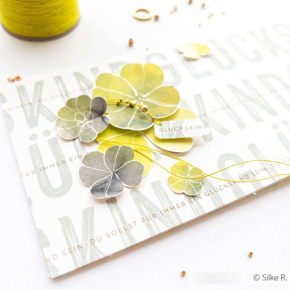 Karte mit geschichteten grünen Kleeformen-Grünes Glücksklee-Gestöber-von Alexandra Renke, mit gelben Fäden und Perlen auf Weiß. Ideal für Papierprojekte oder als Glücksbringer für Ihre Bastelarbeiten mit eleganten Akzenten aus Designpapier.