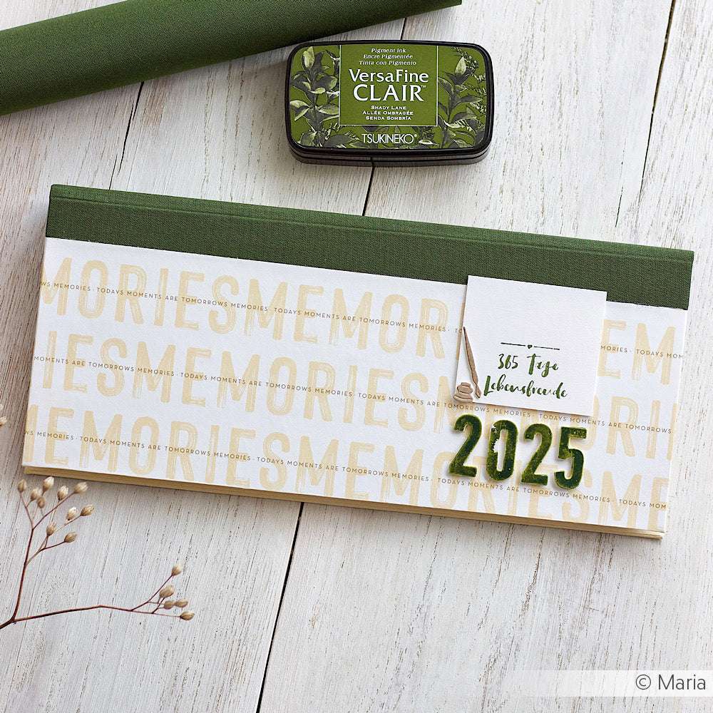 Der grün-weiße Planer 2025 von Alexandra Renke liegt auf einem Holztisch mit grünem Stempelkissen, getrockneten Blumen und Designpapier - ideal für Papierprojekte und Buchbinderei-Liebhaber.