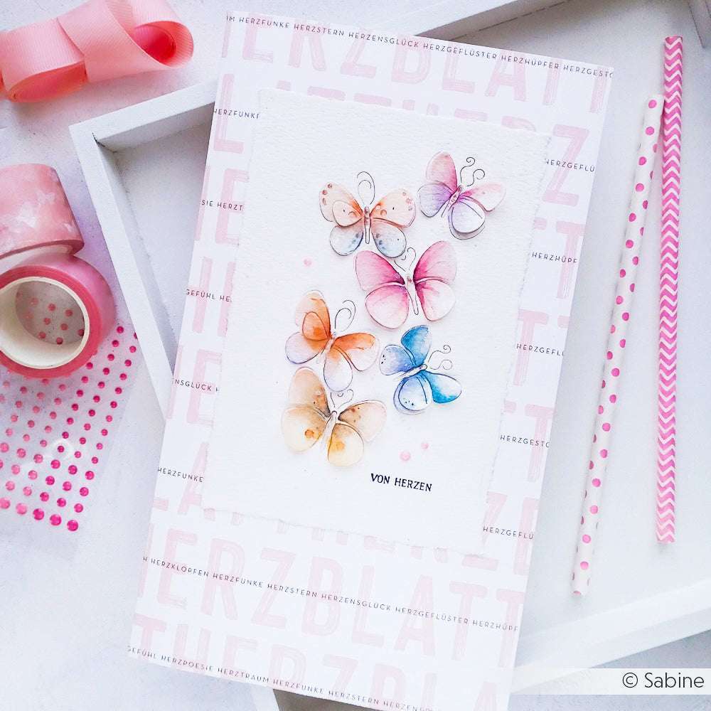 Handgefertigte Karte von Alexandra Renke mit pastellfarbenen Schmetterlingen und "VON HERZEN"-Schriftzug, umgeben von rosafarbenem Bastelmaterial - ideal für Papierprojekte oder zur Kombination mit Ihrem Lieblingsdesignpapier.