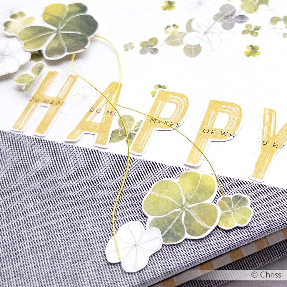 Papierkleeblätter und der Schriftzug "HAPPY" zieren das Designpapier von Alexandra Renke auf grauem Stoff - eine perfekte Inspiration für Papierprojekte oder Buchbinderei.