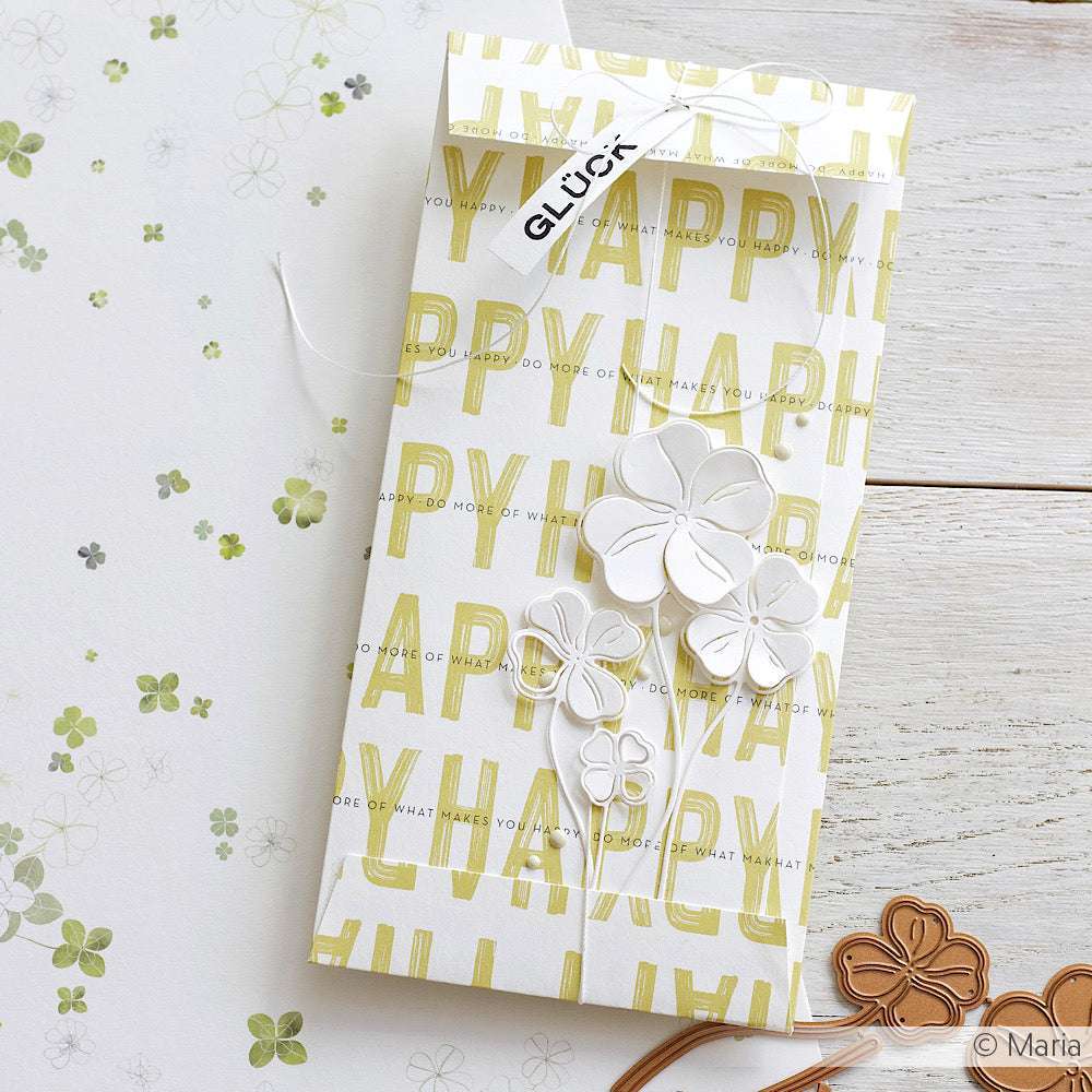Ein verzierter Umschlag von Alexandra Renke mit "HAPPY"-Text, weißen Papierblumen und einem "GLÜCK"-Anhänger auf hellem Holz - ideal für Papierprojekte oder Grußkarten mit stylischem Designpapier.
