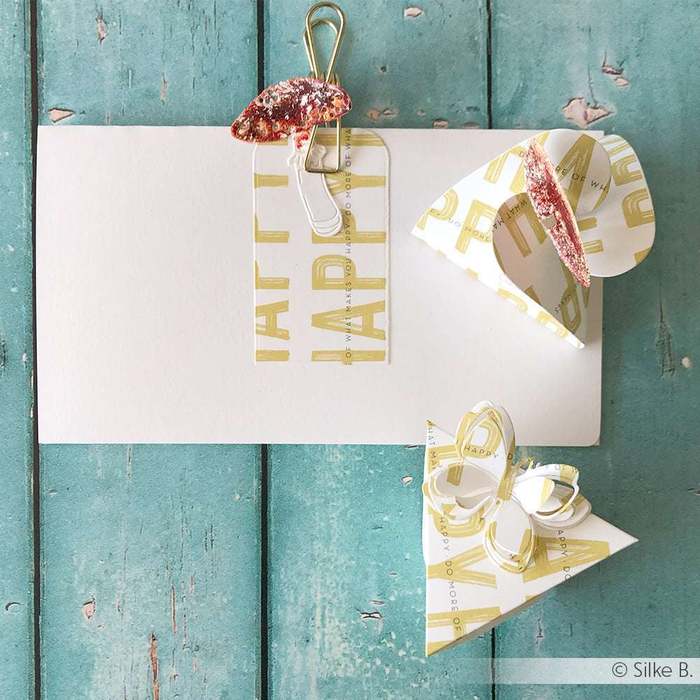 Handgefertigte Papierprojekte von Alexandra Renke und eine HAPPY-Karte auf einer tealfarbenen Holzoberfläche mit einer Büroklammer - ideal für die Buchbinderei oder als Inspiration für kreatives Designpapier.