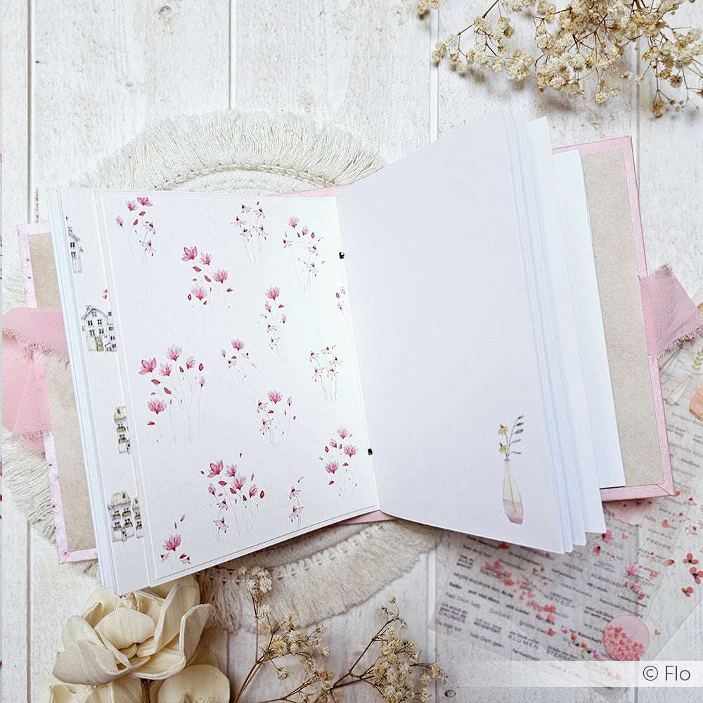 Aufgeschlagenes Notizbuch von Alexandra Renke mit rosafarbenen Blumenillustrationen auf weißen Seiten, ideal für Papierprojekte oder das Erstellen von Grußkarten, ausgestellt zwischen Trockenblumen auf einem weißen Holztisch.