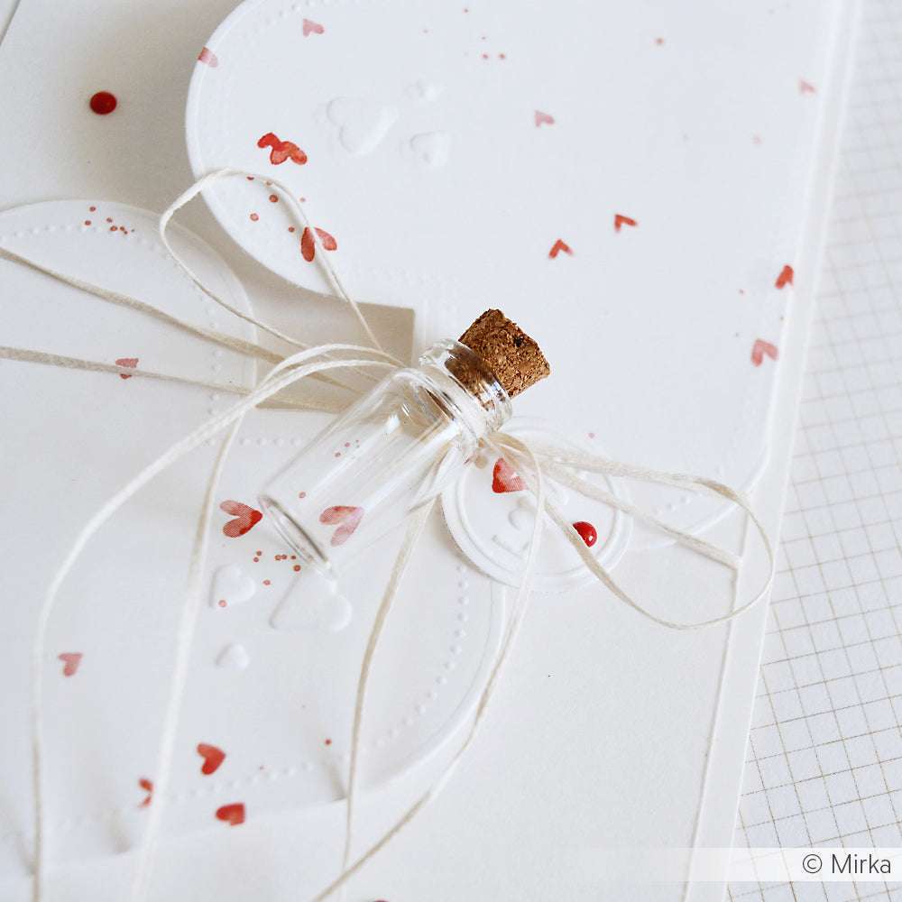 Eine kleine Glasflasche mit Korken sitzt auf einer weißen Karte von Alexandra Renke, die mit roten Herzen und einer gebundenen Schnur verziert ist - ideal für einzigartige Papierprojekte oder personalisierte Grußkarten.