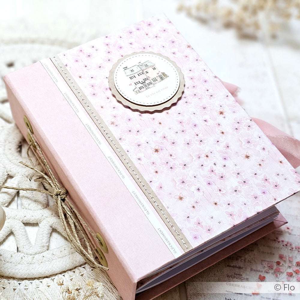 Das Scrapbook-Album von Alexandra Renke aus Blütenmuster rosa Designpapier hat einen dekorativen Einband, ein Bindeband und ein kreisförmiges Etikett mit kleinem Text und Noten - ideal für Papierprojekte.