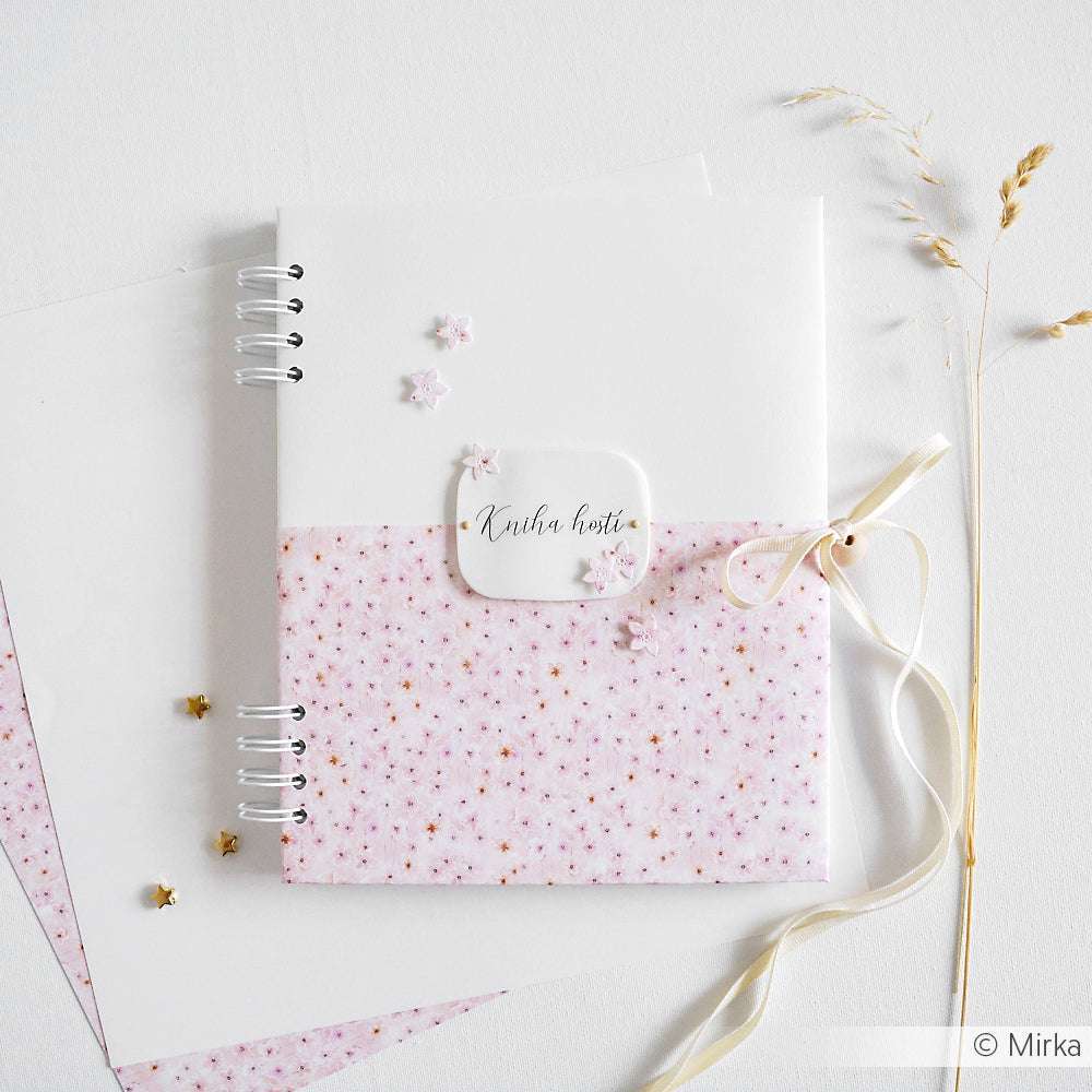Ein Alexandra Renke-Notizbuch mit Blütenmuster rosa, verziert mit einem Band und Sternakzenten, wird auf einer weißen Fläche präsentiert.