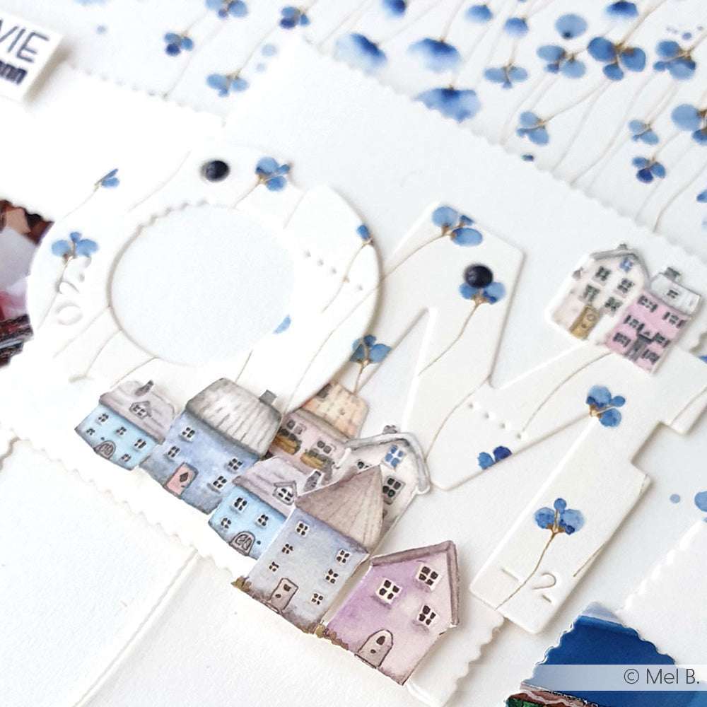 Nahaufnahme der Papierkunst, die HOME buchstabiert, mit pastellfarbenen Häusern und blauen Blumenmustern im Hintergrund, hergestellt mit zarten Details aus Bastelpapier von Alexandra Renke.