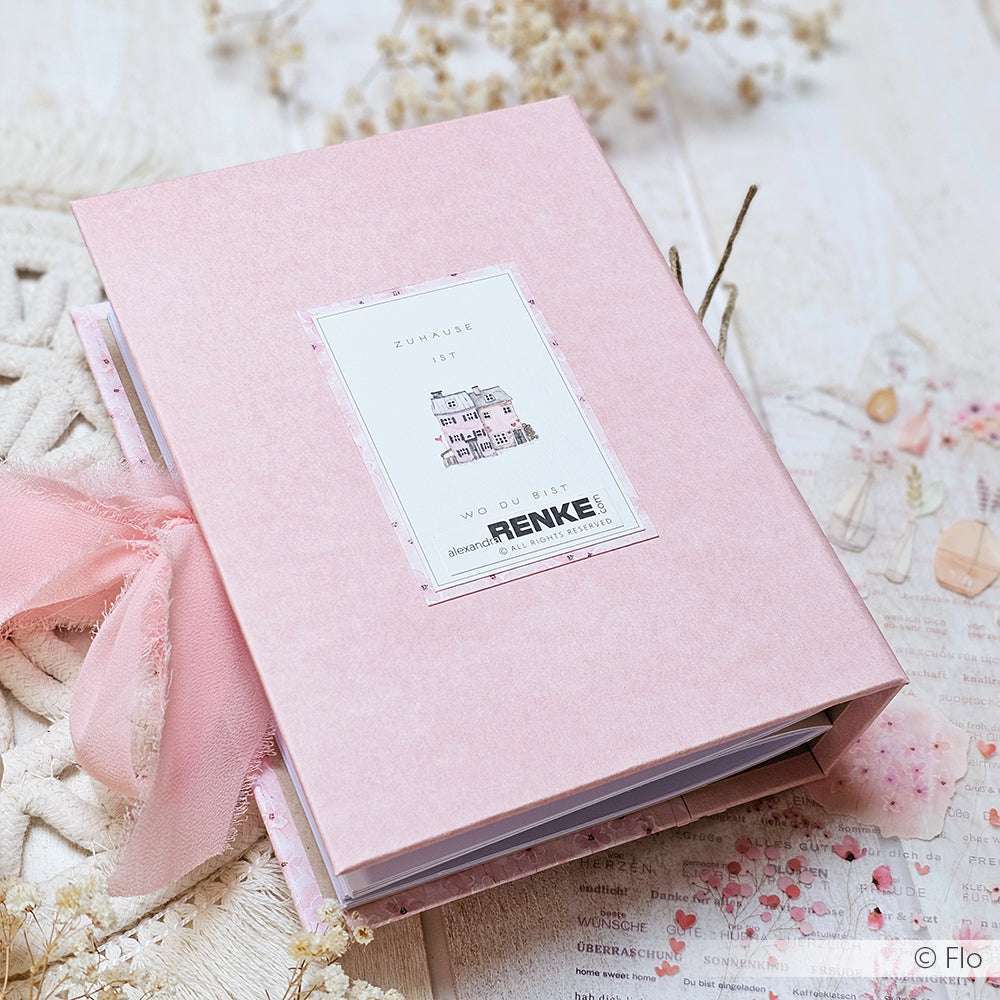 Ein rosafarbenes Scrapbook von Alexandra Renke mit einer Hausillustration auf hochwertigem Designpapier ruht auf einem hellen, dekorativen Hintergrund - eine perfekte Wahl für Papierprojekte und kreative Anwendungen wie den Kärtchenbogen Feine Blütenwelt.