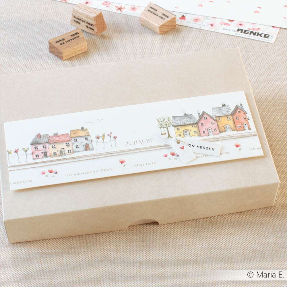 Eine beigefarbene Alexandra Renke Geschenkbox mit einem Etikett mit bunten Häusern und deutschem Text, umgeben von Gummistempeln - ideal für Papierprojekte oder um Ihrem nächsten Buchbinderei-Projekt kreatives Flair zu verleihen.
