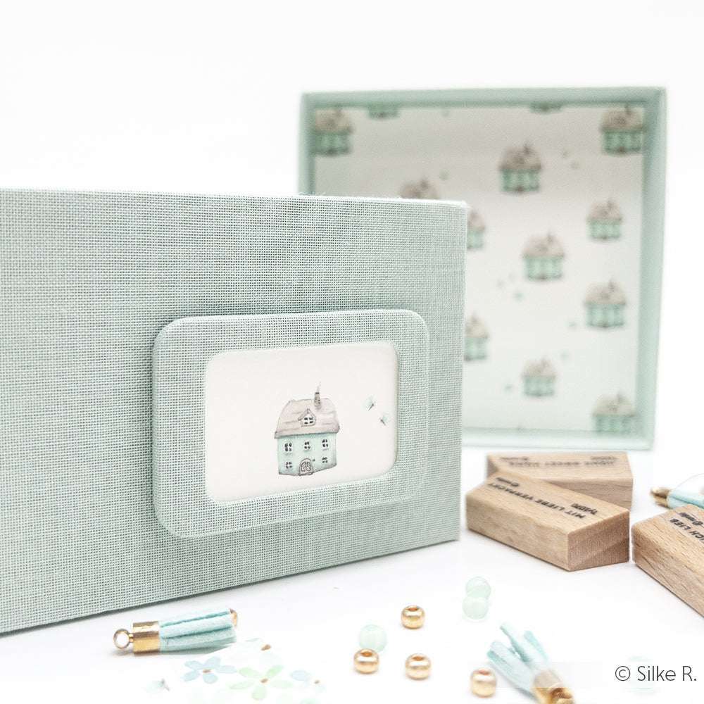 Hellblaue strukturierte Geschenkbox von Alexandra Renke mit einer kleinen Haus-Illustration - ideal zur Aufbewahrung von Designpapier oder Zubehör für Buchbinderei und Grußkarten. Offene Schachtel mit Bastelmaterial im Hintergrund.