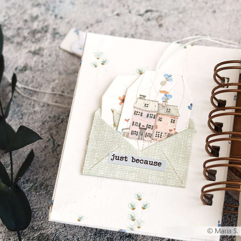 Ein von Alexandra Renke handgefertigter Anhänger mit einem Hausmotiv aus Designpapier wird in einem grünen Umschlag mit der Aufschrift "just because" auf einem aufgeschlagenen Spiralheft präsentiert.