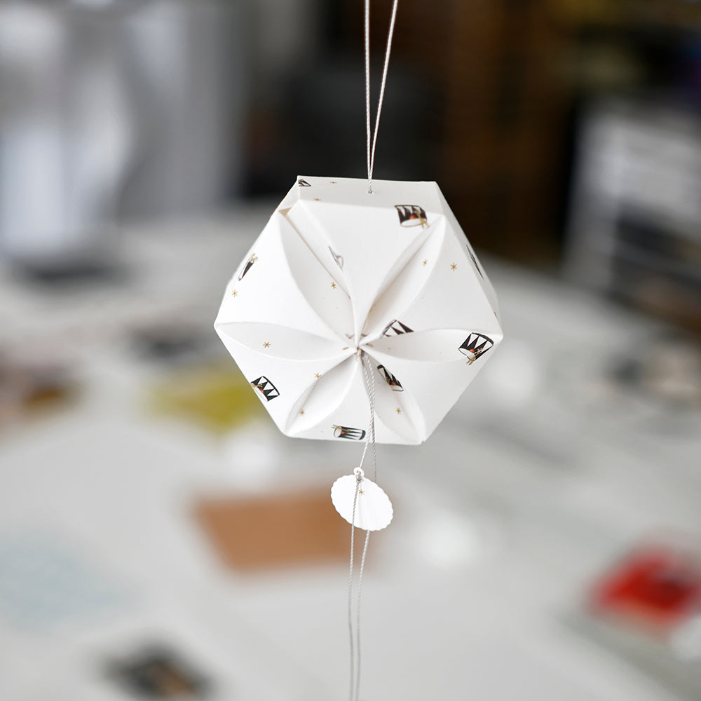 Ein geometrisches, weißes Papierornament aus Designpapier von Alexandra Renke mit verschlungenen Mustern hängt an einer Schnur mit verschwommenem Hintergrund.