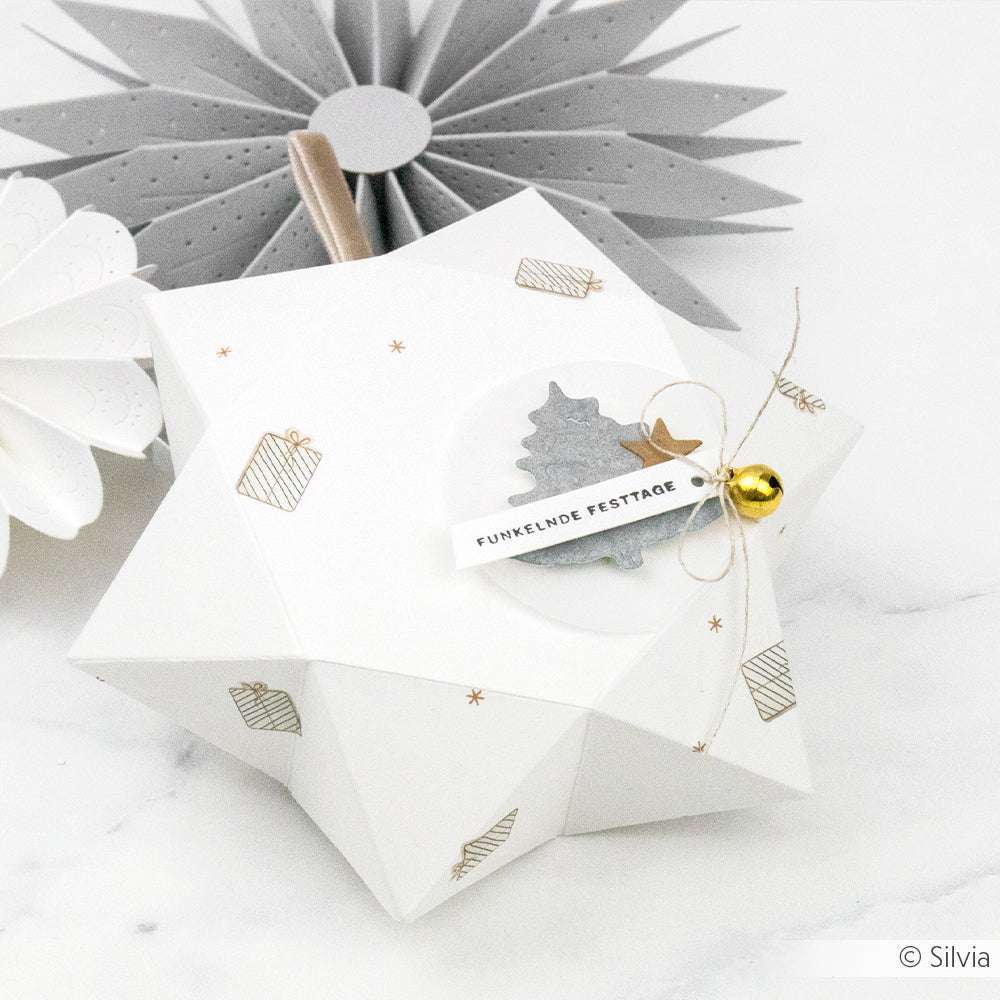 Eine weiße, sternförmige Geschenkbox von Alexandra Renke mit festlichen Anhängern, einer Glocke und goldenen Geschenkmotiven auf Designpapier ruht auf Marmor - ideal für Papierprojekte oder kreative Grußkarten.